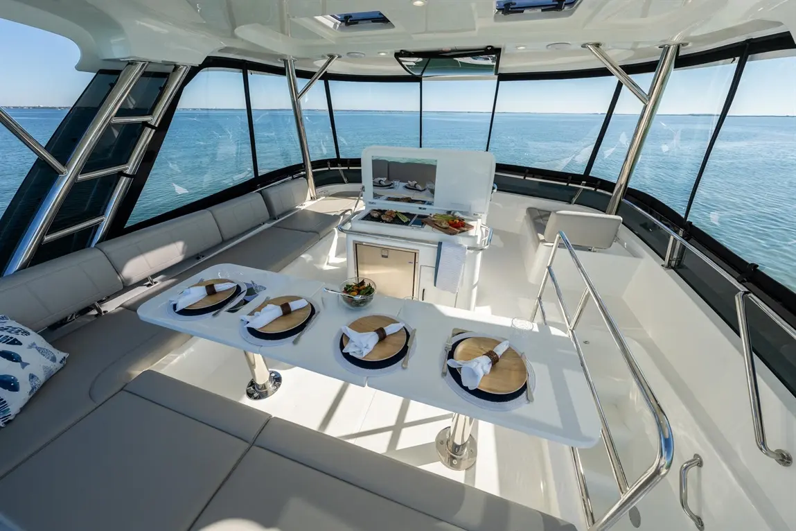 A luxury yacht interior featuring a dining table set for four, with a view of the sea and a cooking area in the background.